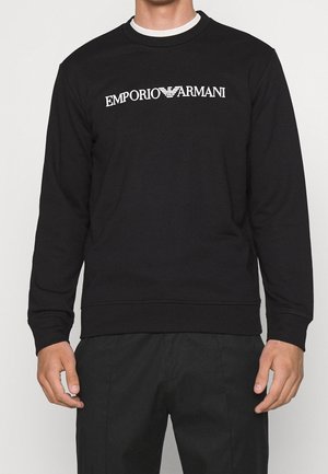Mies, jolla musta pitkähihainen collegepaita, jossa valkoinen ”EMPORIO ARMANI” -logo rinnan keskellä, ja mustat housut.