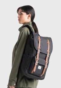 Svart ryggsäck med bruna läderremmar, grått tyg, en framficka och silverfärgade metallbeslag. Slät textur och rektangulär form.