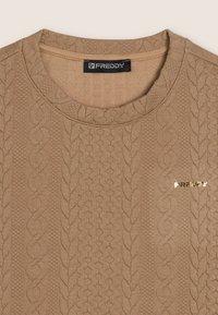 Suéter de punto color coñac con patrones de trenzas y textura acanalada. Presenta un escote redondo y un pequeño logo dorado en el lado izquierdo.