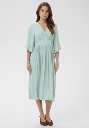 Femme portant une robe midi bleu clair à col en V et manches trois-quarts, associée à des chaussures plates tissées marron, debout sur un fond blanc.