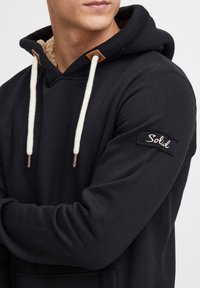 Sudadera negra con un interior suave forrado de felpa, que presenta cordones blancos y un detalle de cuero en la capucha. "Solid" bordado en la manga.