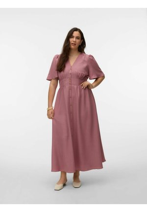 Kvinne iført en støvrosa maxikjole med knapper, korte puffermede ermer og beige sko med lave hæler, stående med en hånd på hoften.