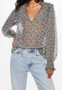 Chemisier noir transparent avec imprimé floral blanc, manches longues et poignets à volants. Associé à un jean bleu clair taille haute.