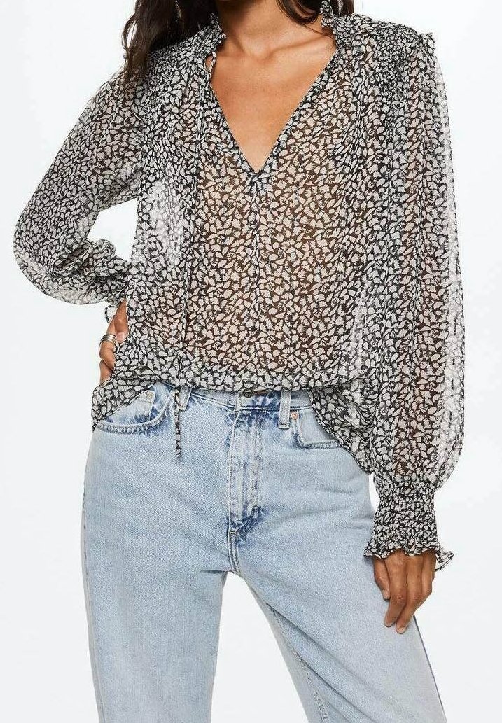 Chemisier noir transparent avec imprimé floral blanc, manches longues et poignets à volants. Associé à un jean bleu clair taille haute.