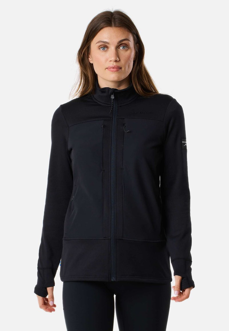 Femme aux longs cheveux bruns portant une veste zippée noire avec des ouvertures pour les pouces, se tenant devant un fond clair et uni.