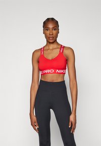 Sujeción deportiva roja con tirantes anchos que presenta el logo "NIKE PRO", combinada con leggings negros de cintura alta, de corte estándar y textura suave.