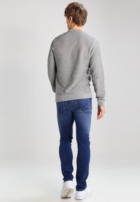 Hombre caminando alejándose, vestido con un suéter gris, jeans azules y zapatillas blancas, contra un fondo claro y simple.