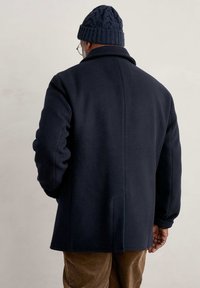 Cappotto di lana blu navy con colletto strutturato e maniche rilassate, abbinato a un berretto in maglia scura. Tessuto texturizzato con dettagli minimi.