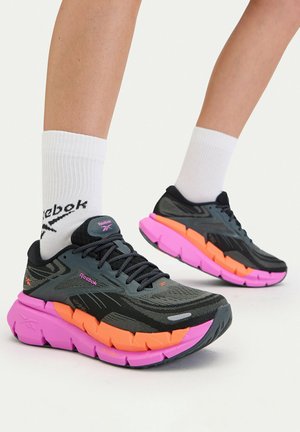Osoba nosząca czarne buty sportowe z pomarańczowymi i różowymi podeszwami oraz białe skarpety Reebok, stojąca na białym tle.