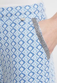 Modré a bílé kalhoty s geometrickým vzorem, které mají kovový lem podél kapsy pro větší detail.