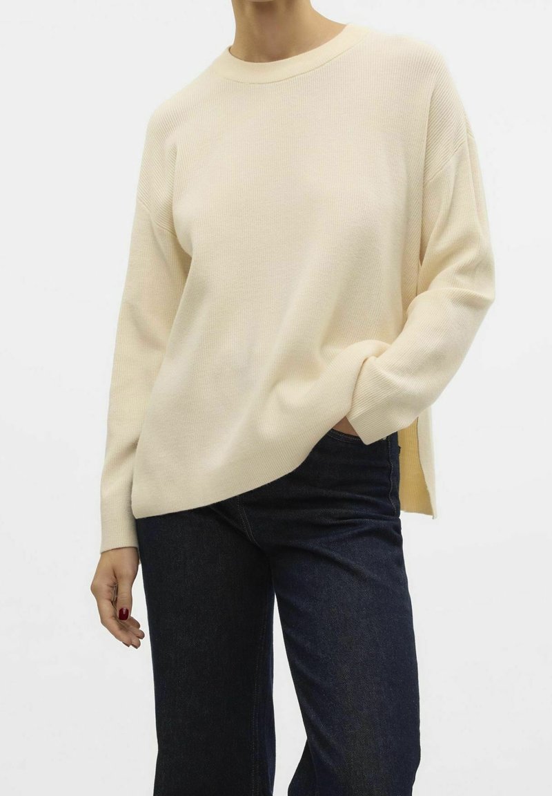 Personne portant un pull côtelé crème à manches longues avec un col rond et un jean bleu foncé, main dans la poche, sur un fond uni.
