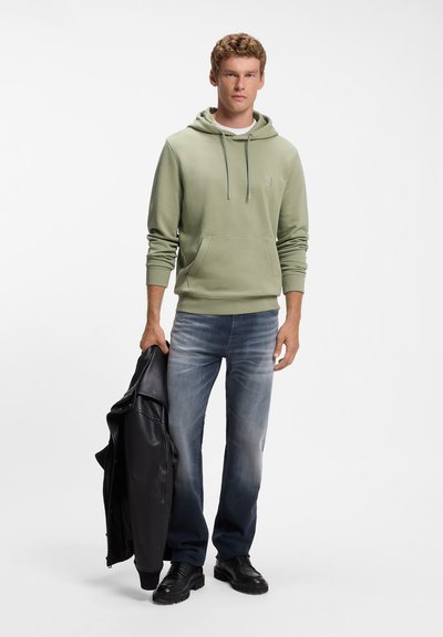 Jeune homme portant un sweat à capuche vert clair et un jean bleu délavé, tenant une veste en cuir noire dans une main, debout sur fond blanc.