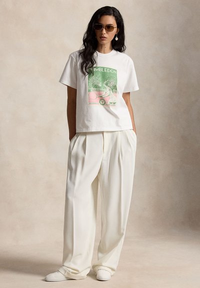 Γυναίκα που φοράει λευκό μπλουζάκι με oversized γραφικό Wimbledon, λευκό παντελόνι με φαρδιά μπατζάκια, λευκά αθλητικά, γυαλιά ηλίου και σκουλαρίκια σε σχήμα δακρύων, όρθια.