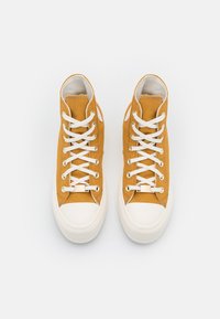 High-top sneakers in mustard yellow canvas, featuring white laces and rubber soles. Double eyelets and subtle stitching details are visible.