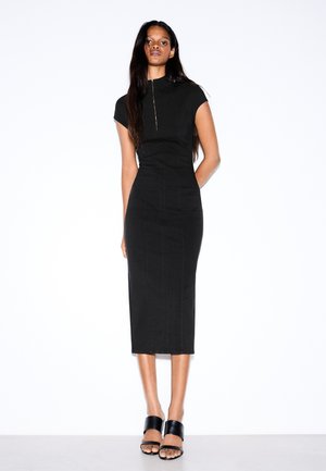 Femme debout portant une robe midi noire ajustée avec des manches courtes et des sandales à talons noires ouvertes à l’avant, sur fond blanc.