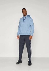 Bărbat stând în picioare, îmbrăcat cu un hanorac albastru deschis și pantaloni de trening negri cu dungi albe pe laterale, purtând adidași negri, pe un fundal simplu.