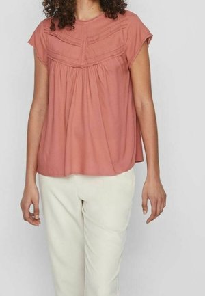 Femme portant un chemisier ample à manches courtes couleur rose poussière avec des détails en dentelle, associé à un pantalon taille haute beige clair.