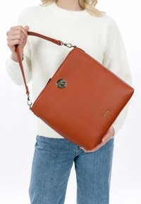 Bolso de cuero marrón con un diseño estructurado, correa larga y un acento metálico redondo. Presenta una superficie texturizada y un branding sutil.
