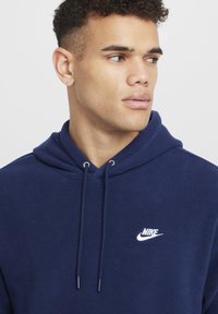 Felpa con cappuccio in pile navy, dotata di cappuccio regolabile e logo Nike bianco sul petto. Texture morbida e design casual, caratterizzata da una tasca a marsupio frontale.