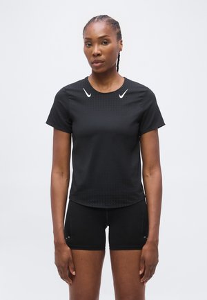Vrouw met gevlochten haar die een zwart sportshirt met korte mouwen en zwarte shorts draagt, staand tegen een effen lichte achtergrond.