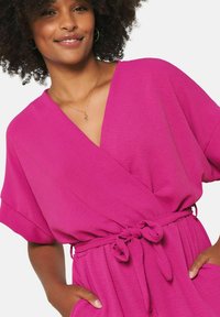 Fuchsia wrap dress with short sleeves, tie waist detail, and textured fabric. Model poses with hands in pockets, showcasing the relaxed fit.