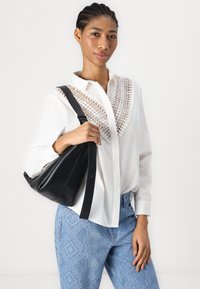 Camicia bianca con chiusura a bottoni e dettagli in pizzo complesso, abbinata a jeans blu con motivo e una borsa a tracolla in pelle nera.