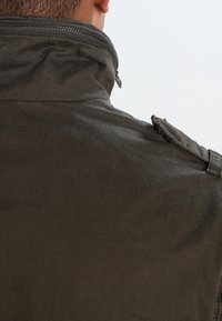 Mørkegrøn jakke med høj krave, udstyret med lynlås og skulderklap. Stoffet virker tekstureret og holdbart.