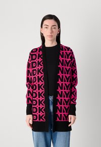 Melnā kardigāna ar drosmīgu rozā atkārtotu "DKNY" rakstu, atvērta priekšpuse, ribinātas aproces un brīvs piegriezums. Uzvilkts virs melna topa un zila džinsu biksēm.