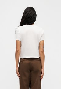 Λευκό κοντό μπλουζάκι με κοντά μανίκια και στρογγυλή λαιμόκοψη, σε συνδυασμό με καφέ παντελόνι που διαθέτει πίσω τσέπες και ραφές.