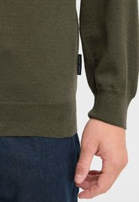 Maglione lavorato a maglia verde oliva con polsini e orlo a costine. Presenta un piccolo etichetta sul lato, indossato sopra pantaloni in denim scuro.