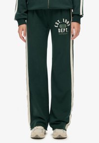 Groene sweatpants met crème zijstrepen, elastische tailleband en bedrukt text "EST. 1982 SD DEPT. SUPERDRY" in het wit. Zachte stoftextuur.