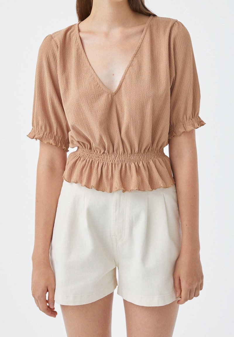 Femme portant un chemisier marron clair texturé à col en V avec des manches bouffantes et un short blanc taille haute, debout devant un fond uni.