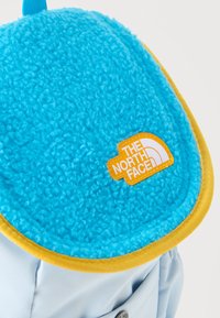 Μπλε φλις πτερύγιο με κίτρινο περίγραμμα σε ανοιχτό μπλε σακίδιο πλάτης με πορτοκαλί και λευκή ετικέτα λογότυπου The North Face.