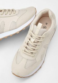 Sneakers atletici beige realizzati in rete e tessuto con una finitura testurizzata, dotati di un design con lacci, suola in gomma e un marchio discreto.