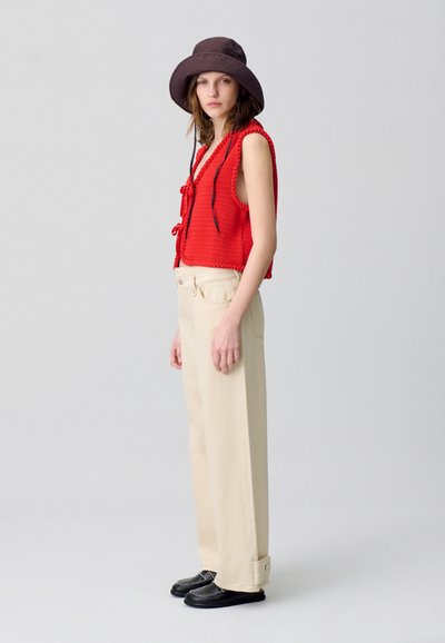Chaleco de ganchillo rojo con lazada frontal, pantalones de pierna ancha beige y zapatos negros. El modelo también lleva un sombrero de cubo marrón. Diseño simple y texturizado.