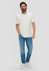 Witte katoenen t-shirt, lichtblauwe denim jeans en witte sneakers. Model poseert met hand in de zak tegen een neutrale achtergrond.