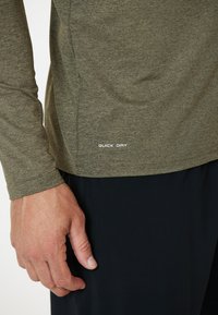 Olivgrünes, langärmliges Sportshirt mit glatter Textur; mit weißem Text "QUICK DRY" nahe dem Saum, kombiniert mit dunklen Hosen.