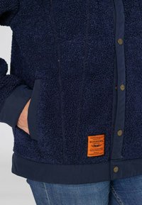 Granatowa polarowa kurtka z teksturowanym wykończeniem, kontrastowymi mankietami z tkaniny, bocznymi kieszeniami oraz pomarańczową metką marki na dolnym hemie.