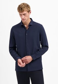 Man met kort krullend haar in een marineblauw lange mouwen overhemd met knopen, past zijn manchet aan met beide handen, witte achtergrond.