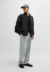 Black lightweight jacket over a textured black sweater, paired with light gray wide-leg pants and black footwear. Gray cap completes the look.