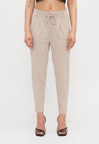 Pantalons beiges fuselés avec taille à cordon, dotés de plis à l'avant et d'une texture lisse. Associés à des escarpins noirs à talons fins.