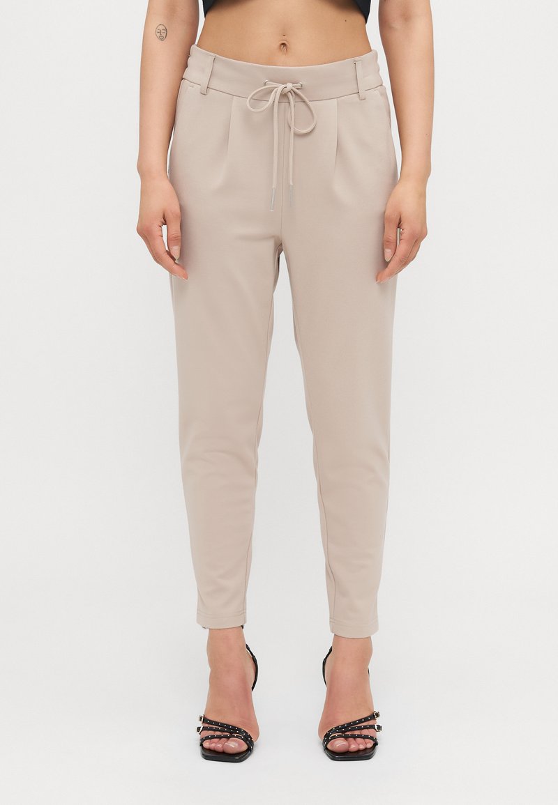 Pantalons beiges fuselés avec taille à cordon, dotés de plis à l'avant et d'une texture lisse. Associés à des escarpins noirs à talons fins.