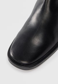 Stivale alla caviglia in pelle nera con una texture liscia, punta arrotondata e suola robusta. Design minimalista senza hardware o dettagli visibili.