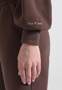 Rękaw brązowej bluzy z ribbowanym mankietem, z haftowanym napisem "Łatwy do pokochania!" w złotym kolorze. Tkanina wydaje się miękka i teksturowana.