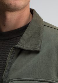 Olijfgroene jas met een ritssluiting, een kraag en een zwart gebreid vest eronder, met een gestructureerde stof en zichtbare naden.