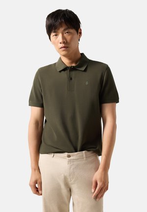 Mężczyzna ubrany w oliwkową koszulkę polo z krótkim rękawem i beżowe spodnie, stoi na tle jednolitego jasnego tła, patrząc przed siebie.