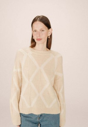 Pull en tricot beige avec un motif en diamant, col et ourlet côtelés, texture douce, associé à un jean bleu clair.