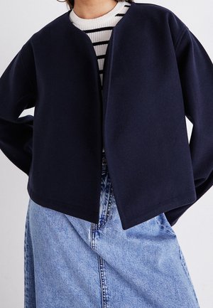 Personne portant une veste bleu marine ouverte sur un pull à rayures blanches et noires, associée à une jupe en denim bleu clair taille haute.