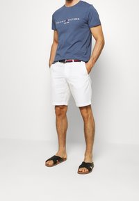 Pánské modré bavlněné tričko s logem "Tommy Hilfiger", bílé bavlněné šortky a černé sandály s křížovými pásky, stojící ležérně s rukama v kapsách.