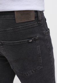 Černé džíny z denimu s hladkým povrchem. Mají hnědou koženou nášivku s označením "MUSTANG" a malou logovou cedulkou na zadní kapse.